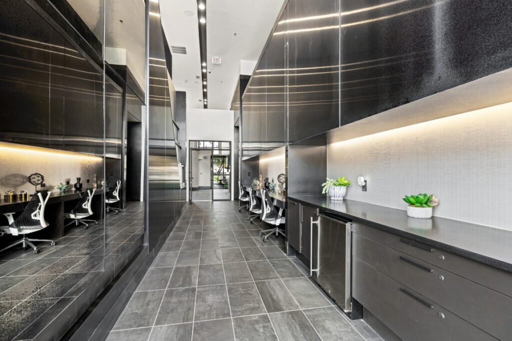 Modern office hallway with black cabinets, gray tile floor, workstations with chairs on one side, reflective wall on the other, and small potted plants on the counters. Natural light enters from glass doors at the end.