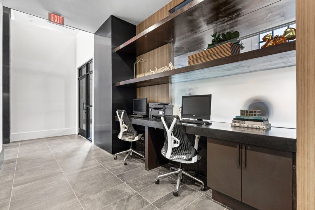 Modern office nook with two computer workstations, black countertops, ergonomic chairs, open wooden shelves with decor items, printer, and books. Gray tile floor and glass door are visible on the left side.