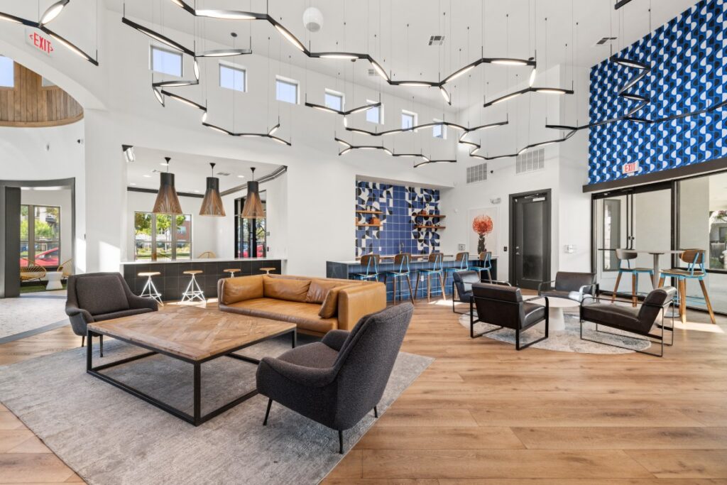 Modern lounge with tan leather sofa, black chairs, wooden table, blue accent wall, barstools, artistic lighting, large windows, and wood floors in a bright, open space.