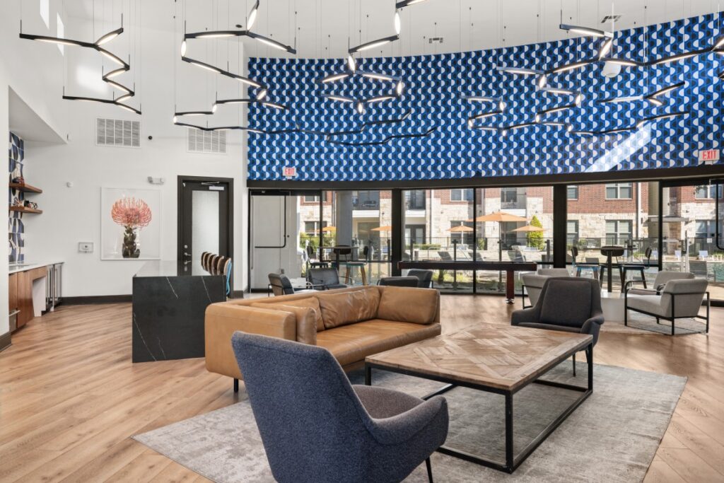 Modern lounge with tan leather sofa, gray chairs, wood floors, geometric blue accent wall, unique ceiling lights, and large windows showing an outdoor patio and apartment buildings.