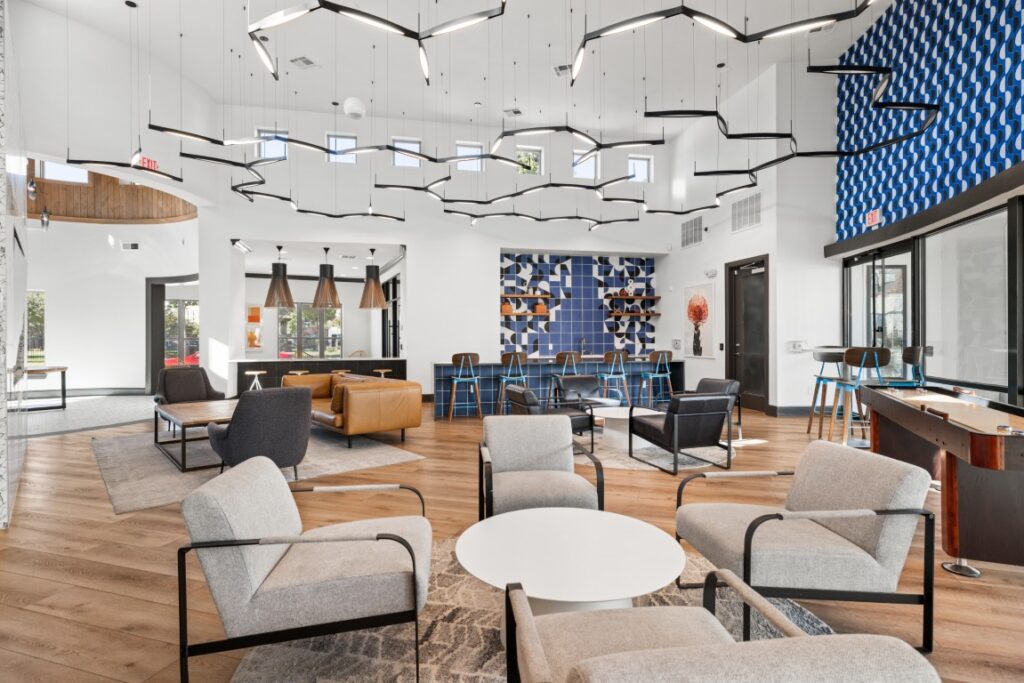 Modern lounge with gray armchairs, round white tables, leather seating, geometric lighting, wooden floors, and a blue accent wall with shelves. Large windows let in natural light.