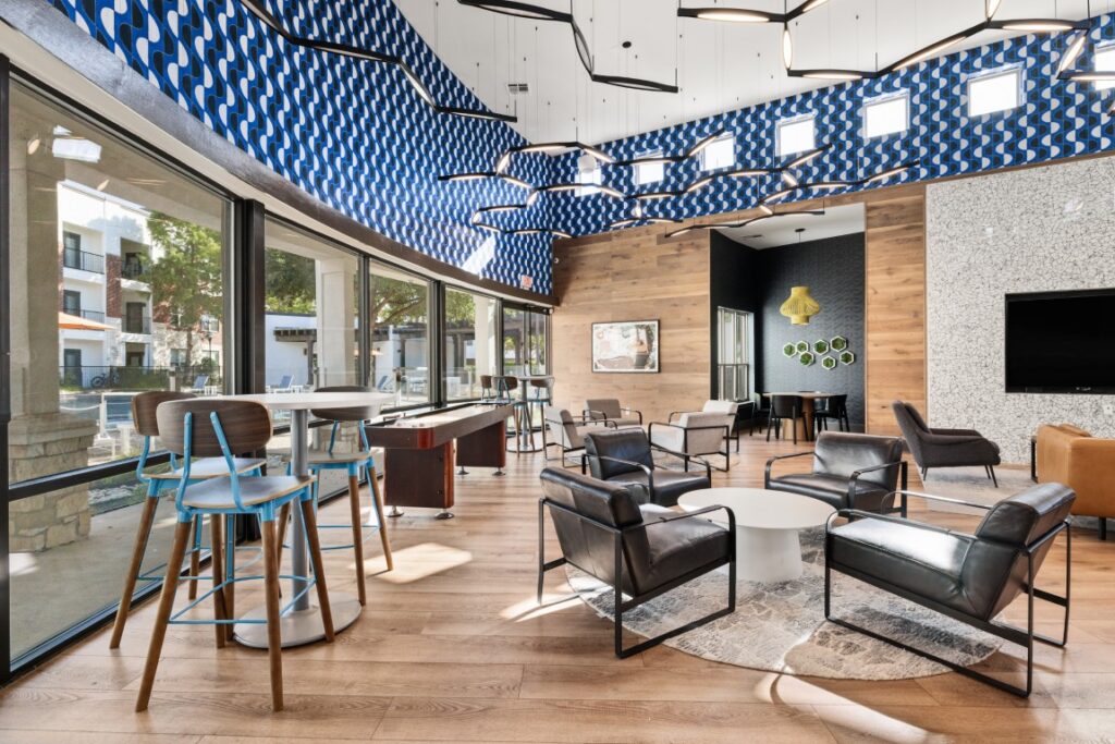 Modern lounge with floor-to-ceiling windows, blue patterned ceiling, black chairs around round tables, barstools at a counter, and a TV on a wall. Natural light fills the space, highlighting wood and stone accents.