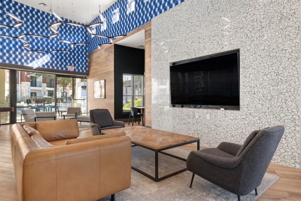 Modern lounge with tan leather sofas, a gray armchair, a large wall-mounted TV, abstract blue ceiling art, wood accents, and large windows letting in natural light.
