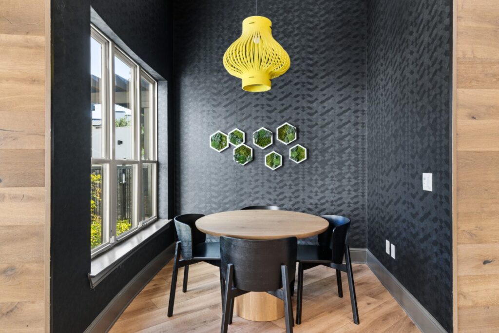 A small dining nook with a round wooden table and four black chairs, a yellow pendant light above, black textured walls, and hexagon-shaped wall planters. Large window lets in natural light.