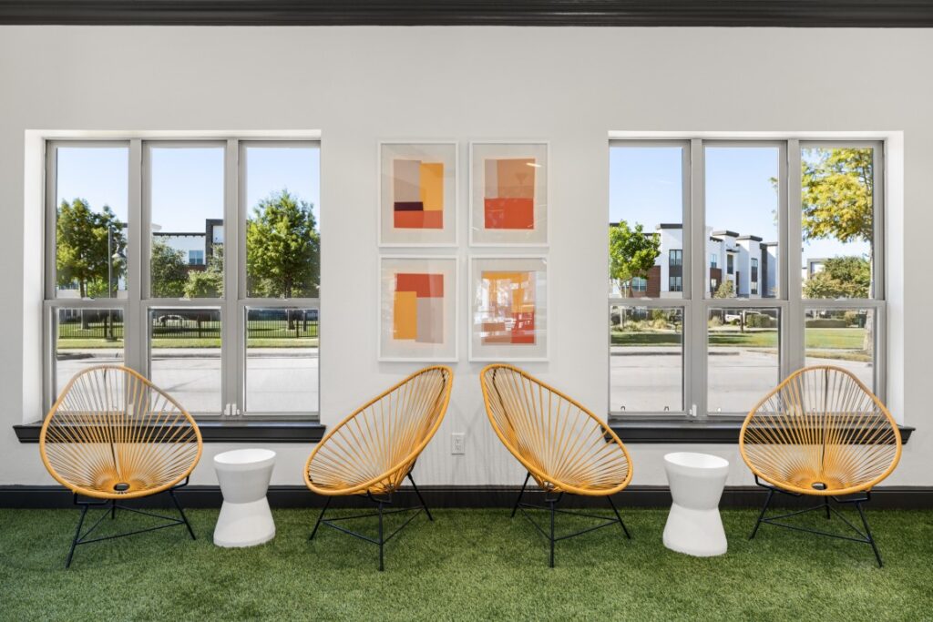 Four modern yellow chairs and three white stools sit on green carpet between two large windows. Four abstract orange and red art pieces hang on the white wall. Trees and buildings are visible outside.