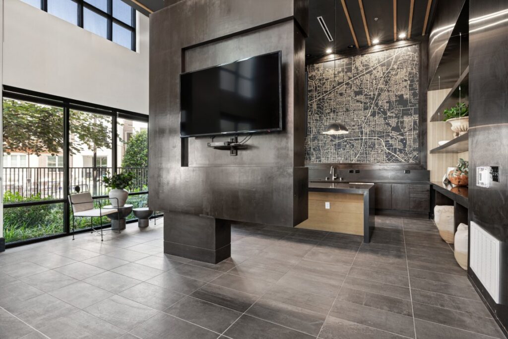 Modern lounge area with large TV, black tiled floors, floor-to-ceiling windows, a wall map, stylish seating, a small kitchen, and greenery visible outside.