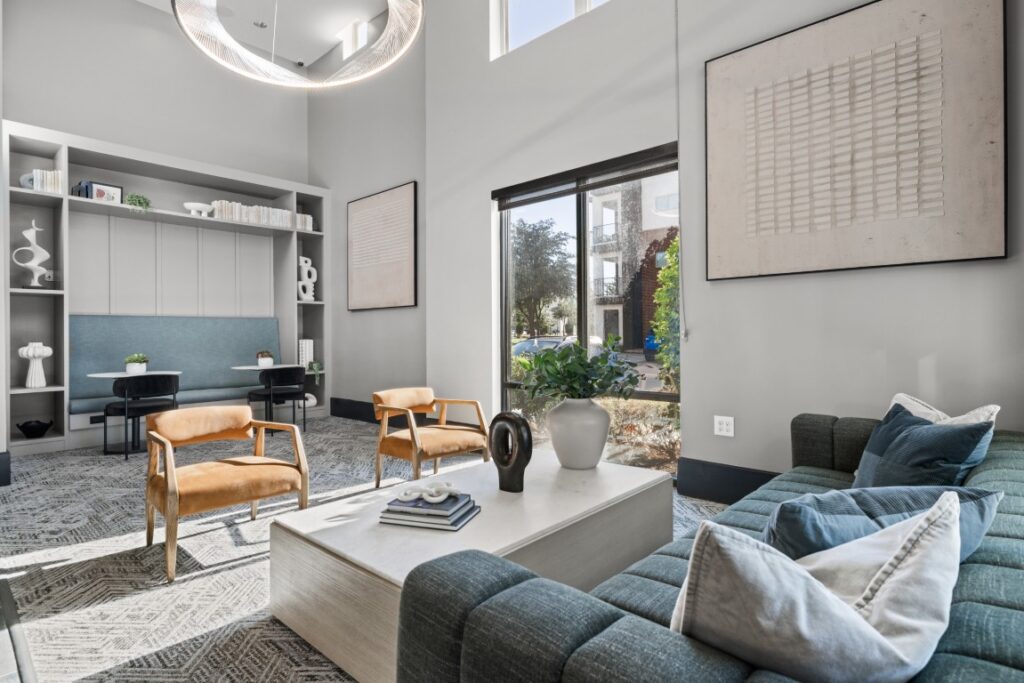Modern living room with blue sofa, two tan chairs, a white coffee table, large wall art, built-in shelves, and a window letting in natural light. A small desk area is visible in the background.