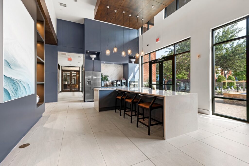Modern lobby with a marble island, four bar stools, pendant lights, large windows, and blue-gray walls. Sunlight fills the space, highlighting minimalist decor and outdoor views.