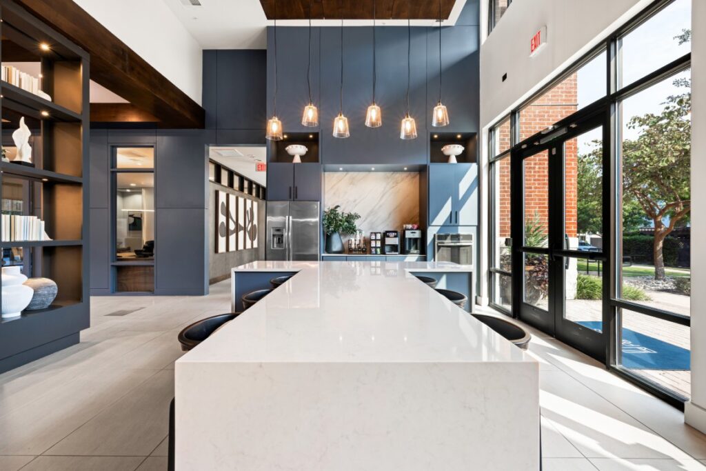 Modern kitchen with a white marble island, bar stools, pendant lights, built-in shelves, stainless steel appliances, large windows, and a view of trees outside. The space is bright and contemporary.