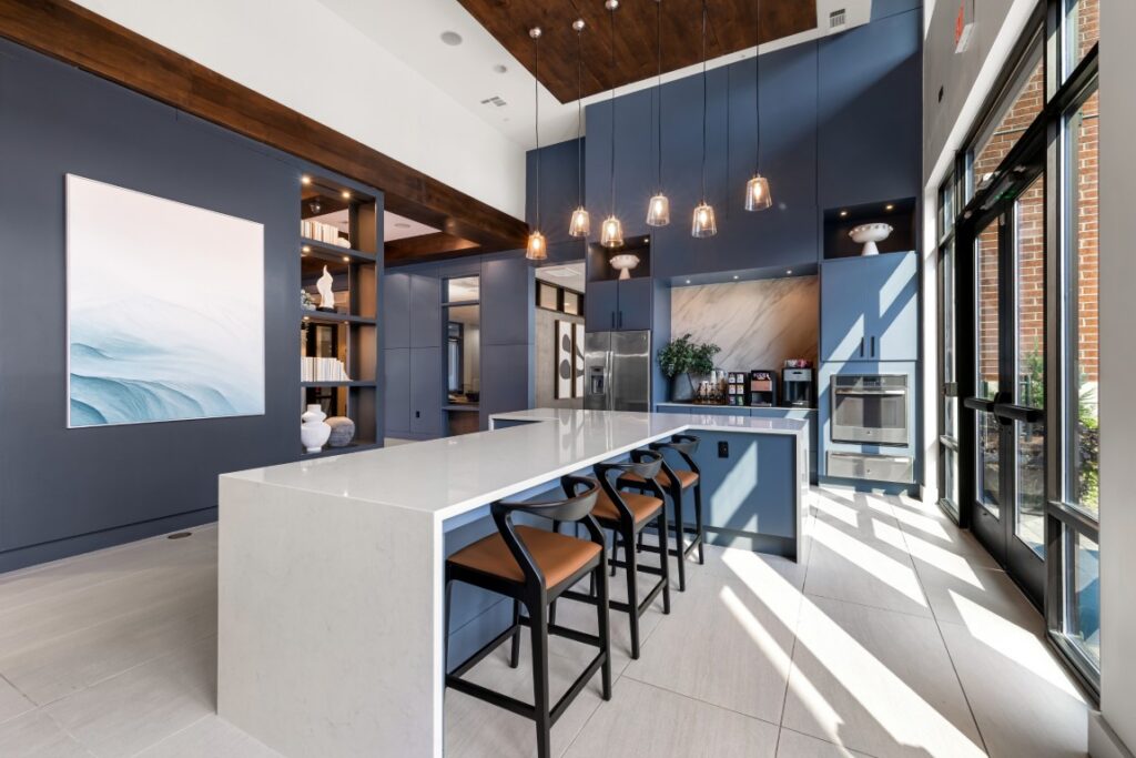 Modern kitchen with blue cabinets, white island, brown barstools, pendant lights, large windows, abstract art, built-in oven, and shelving with decor. Sunlight streams in, highlighting the sleek, contemporary design.