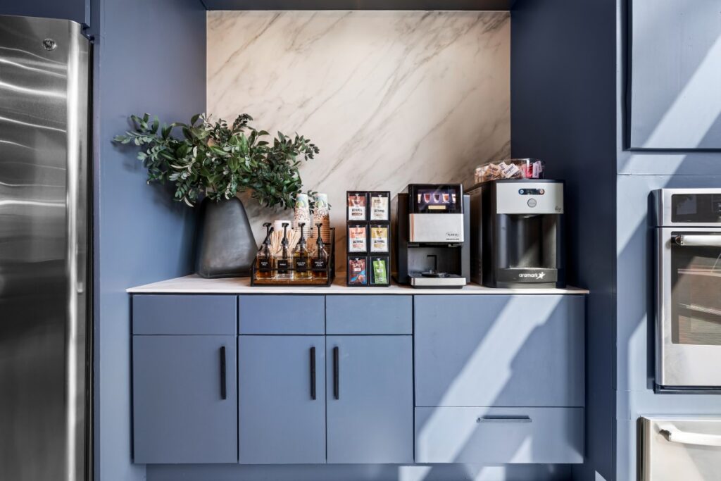 A coffee station with a coffee machine, syrup bottles, tea bags, mugs, a water dispenser, and a vase with greenery on a marble countertop with blue cabinets. Sunlight casts shadows across the setup.