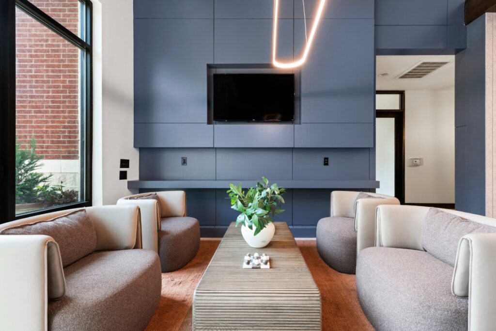 Modern lounge area with four beige armchairs, a wooden coffee table with a plant centerpiece, a wall-mounted TV, blue cabinetry, large window, and unique ceiling light fixture.