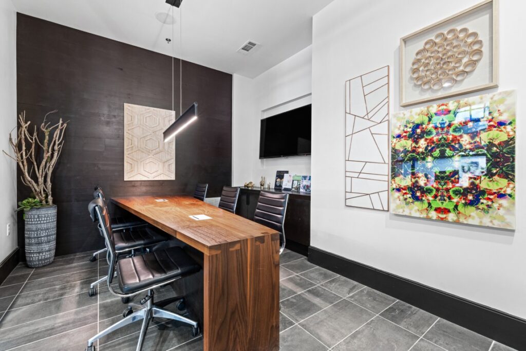 Modern office with a wooden table, four black chairs, wall art, a TV, and a tall vase with branches. The floor is tiled in gray, and the room is bright with white walls and a black accent wall.