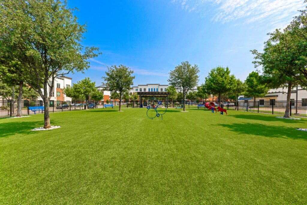 A green, open grassy park with scattered trees, modern playground equipment, and surrounding buildings under a blue sky with light clouds.