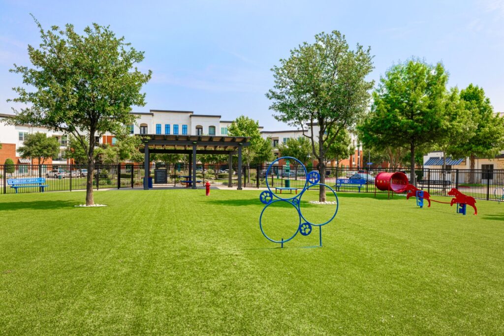 A fenced dog park with green artificial grass, agility equipment like hoops and tunnels, and several trees. Apartment buildings and a pergola are visible in the background under a clear sky.