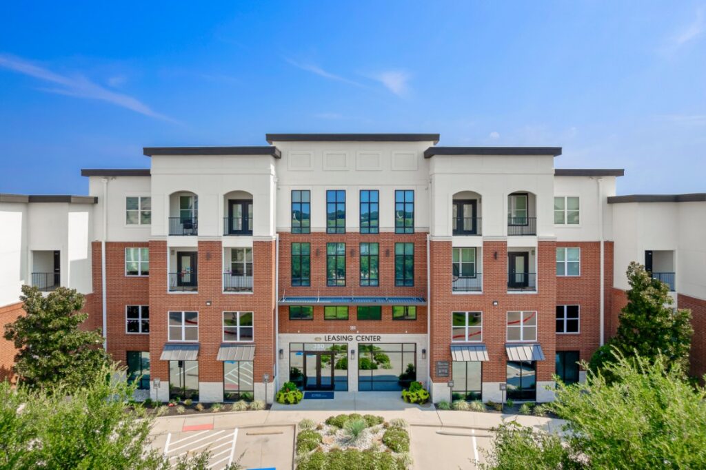 A modern, four-story apartment building with red brick and white siding, large windows, and a Leasing Center entrance. Trees and shrubs are in front, under a clear blue sky.
