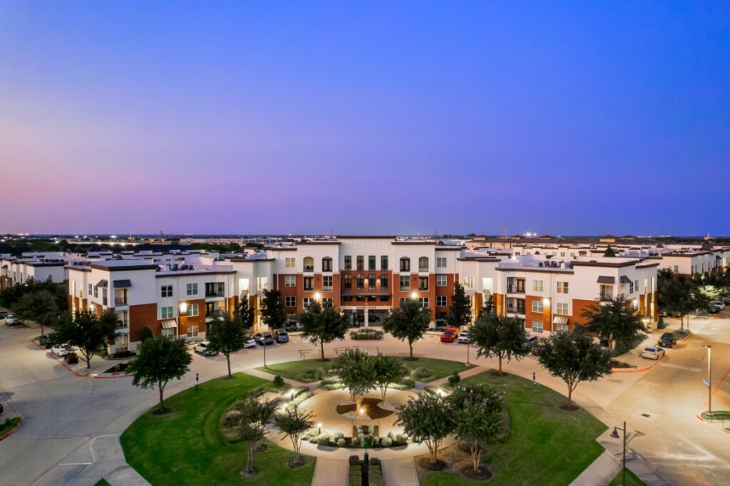 A modern apartment complex with three-story buildings surrounds a circular green space with trees and a central fountain at sunset.