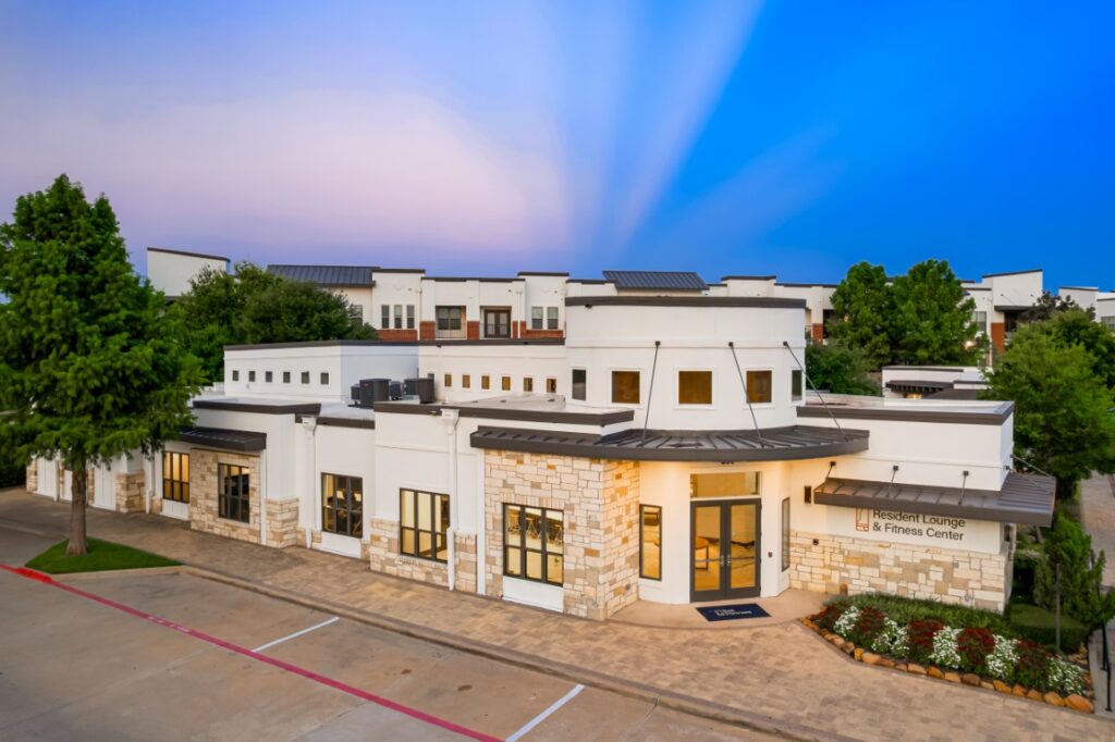 Modern building with white walls, stone accents, large windows, and a sign reading Resident Lounge & Fitness Center, surrounded by trees and landscaping, under a clear blue and purple sky.