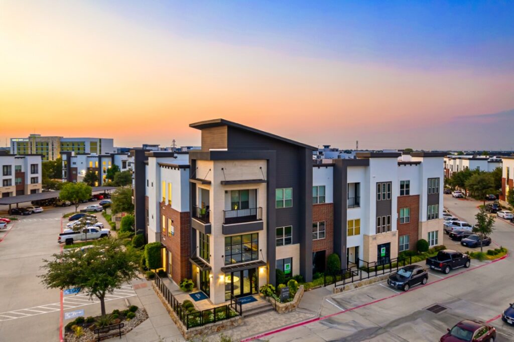 Modern apartment building with large windows and balconies, surrounded by parked cars and greenery, under a colorful sunset sky in a suburban area.