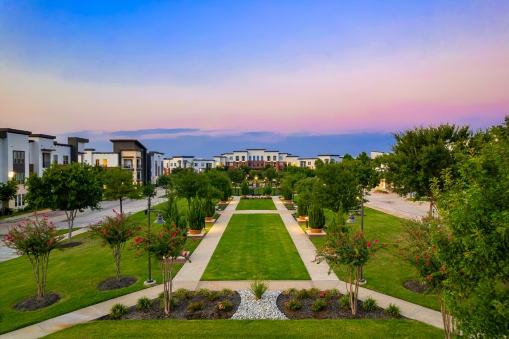 A landscaped walkway with lush green lawns, flower beds, and trees, bordered by modern apartment buildings under a pastel sunset sky.