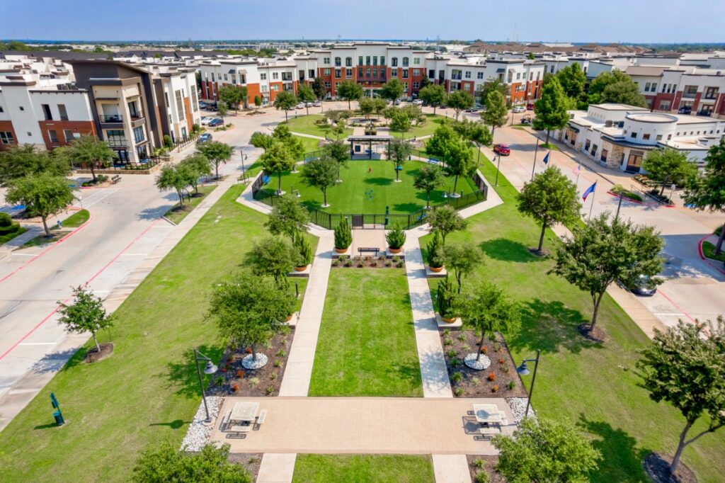 Aerial view of a green park with walkways, trees, benches, and a small pavilion, surrounded by modern apartment buildings and streets on a sunny day.