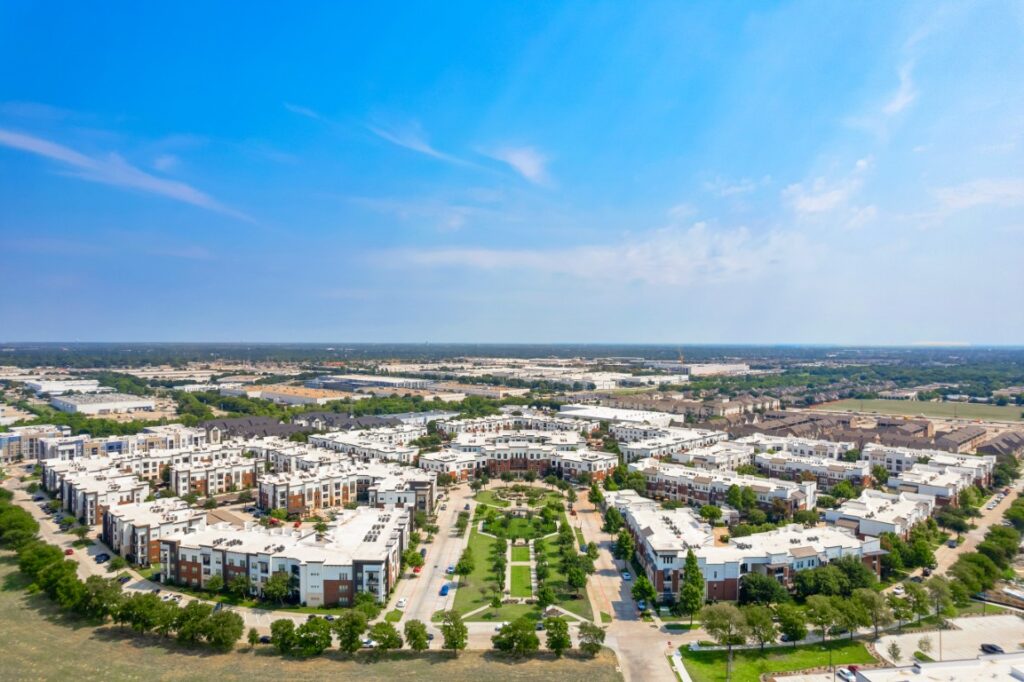 Aerial view of a suburban neighborhood with rows of modern apartment buildings, green spaces, and trees under a blue sky with scattered clouds.