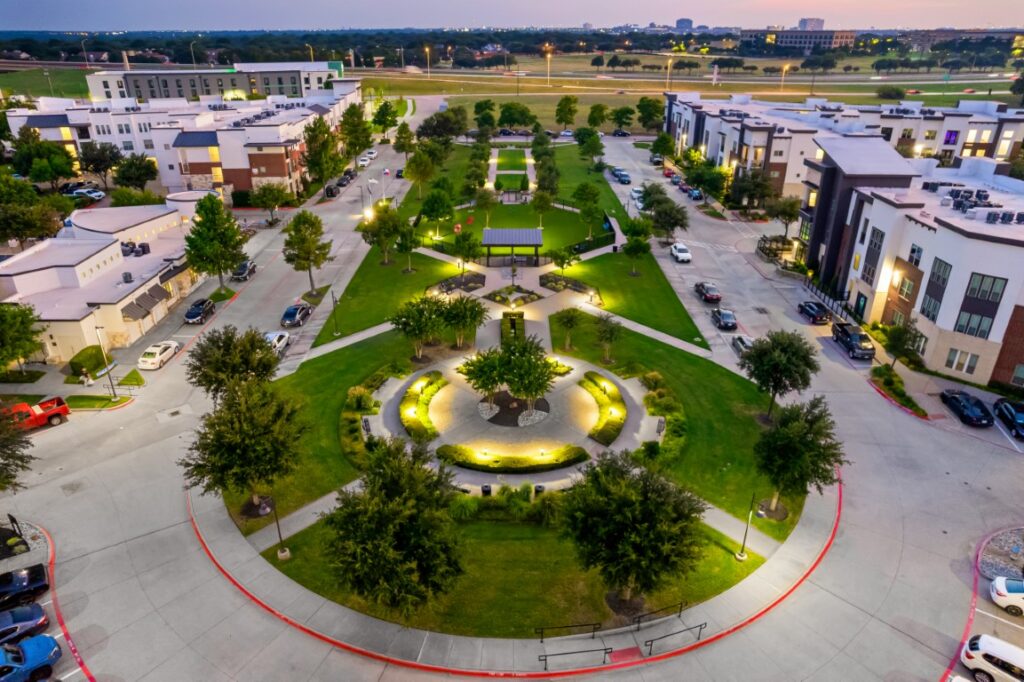 Aerial view of a landscaped urban park with circular pathways, trees, grass, modern buildings, parked cars, and illuminated pathways at dusk.