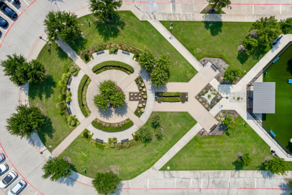 Aerial view of a landscaped park with circular walkways, green lawns, benches, trees, a covered pavilion, and nearby parking spaces on the left.