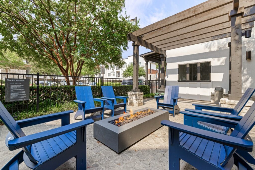 Six blue Adirondack chairs surround a rectangular fire pit under a wooden pergola in an outdoor patio area with trees and a nearby building visible.
