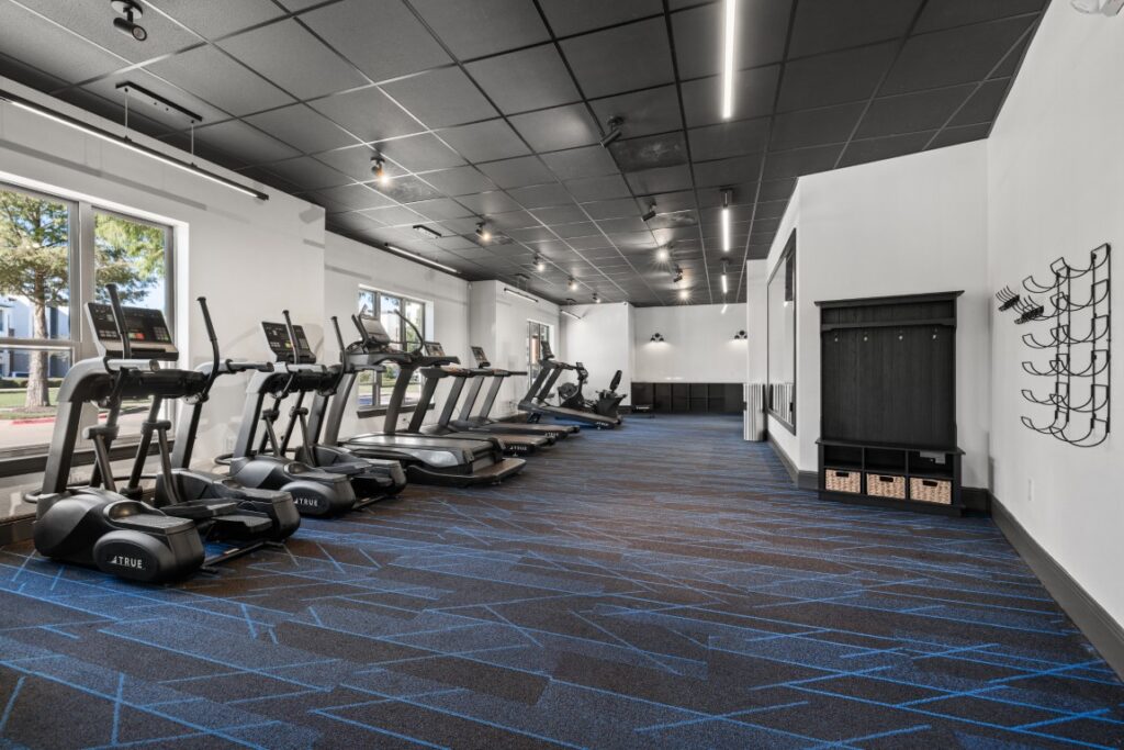 A modern gym with treadmills and elliptical machines lined up by windows, blue-patterned carpet, black ceiling, wall hooks, and a storage bench along the right wall.