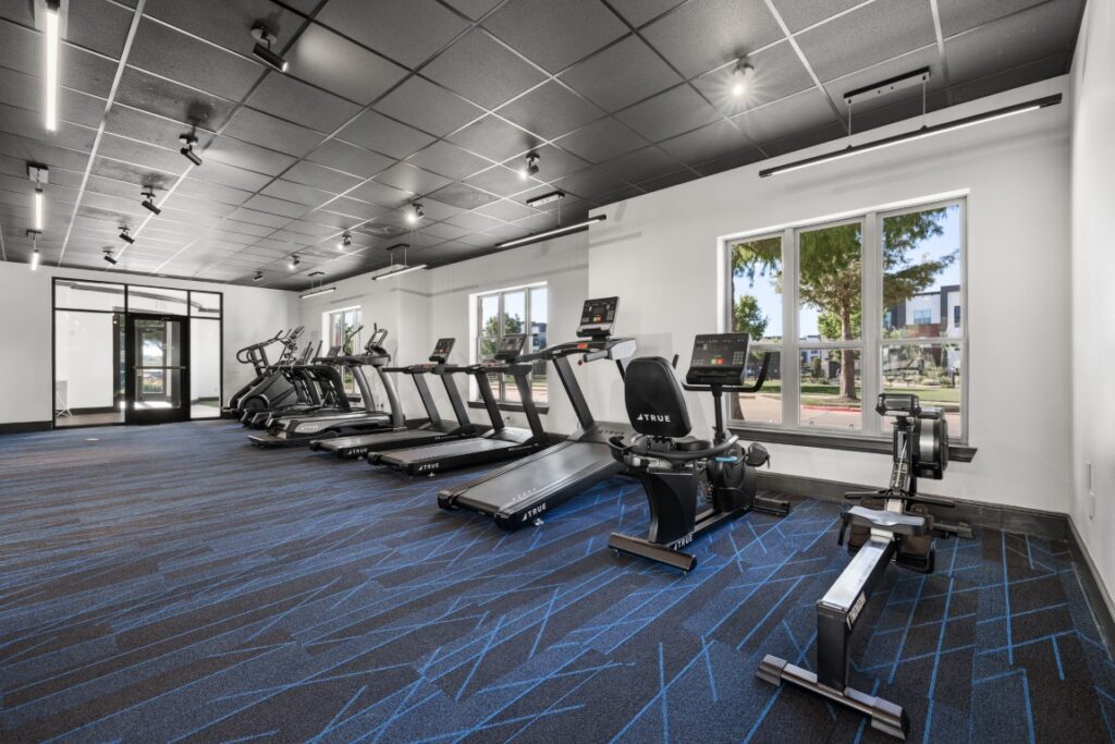 Modern gym with treadmills, stationary bikes, and a rowing machine aligned on blue-patterned carpet, large windows letting in natural light, and black ceiling tiles with track lighting.
