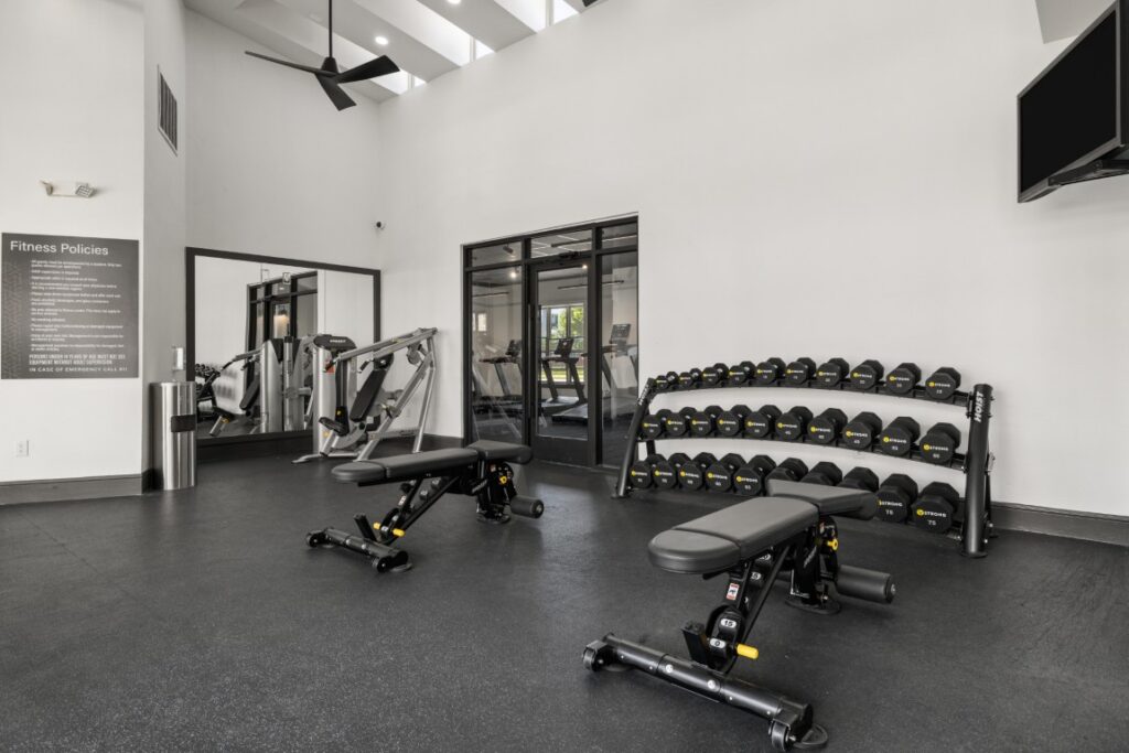 Modern gym with two weight benches, a rack of dumbbells, a cable machine, mirrors, and a wall-mounted TV. The space has white walls, high ceilings, and large black-framed windows. Fitness policies sign is visible.
