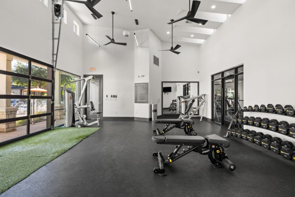 Modern gym with two workout benches, weight machines, and a rack of dumbbells. Large windows, ceiling fans, and artificial turf near the entrance let in plenty of natural light.