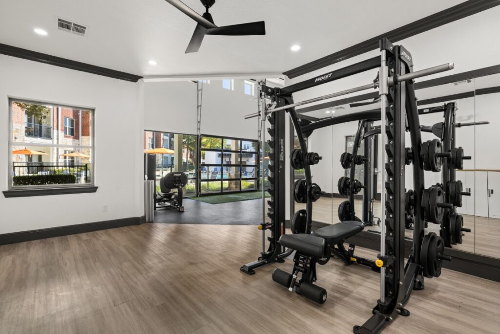 Modern gym with a Smith machine and weight plates on a wood floor, large mirrors, a ceiling fan, and windows showcasing outdoor seating and greenery.