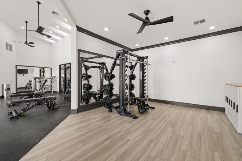 A modern gym with weight machines, barbells, and exercise equipment on black mats, plus wood flooring and ceiling fans in a bright, white room.