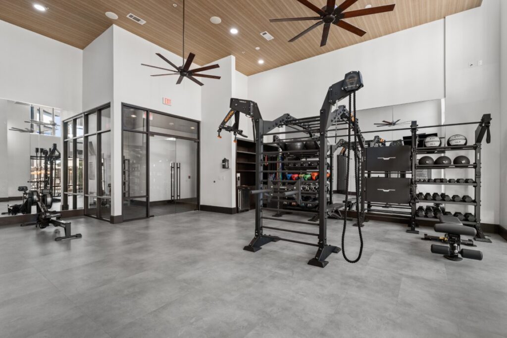 Modern gym with exercise machines, free weights, kettlebells, medicine balls, and benches. The space has large ceiling fans, gray flooring, and white walls with glass doors and windows on one side.