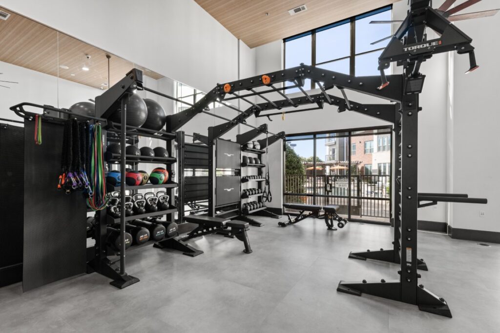 Modern gym with strength training equipment, including a rack with kettlebells, medicine balls, resistance bands, and a large multi-use workout station. Large windows let in daylight and reveal an outdoor view.