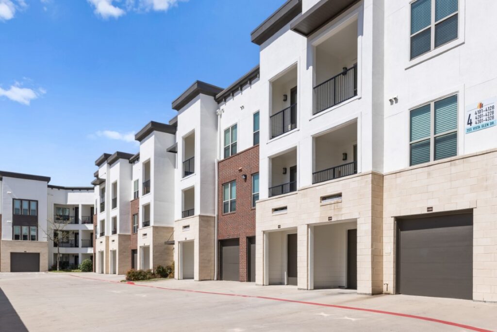 Modern three-story apartment complex with balconies, garages, and a mix of white and brick exterior. The area in front is a wide, clean driveway under a blue sky with some clouds.