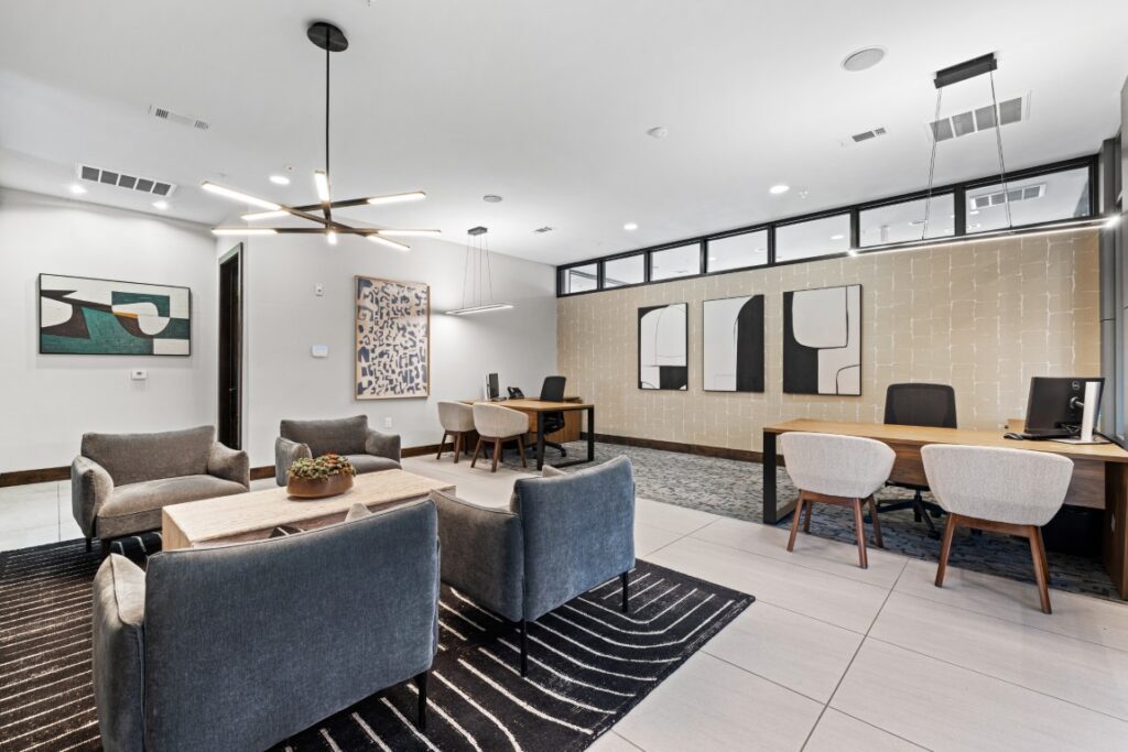 Modern office space with three desks, four gray armchairs, a coffee table with a plant, abstract wall art, large ceiling lights, and a black-and-white rug on tiled flooring. The room is bright and organized.