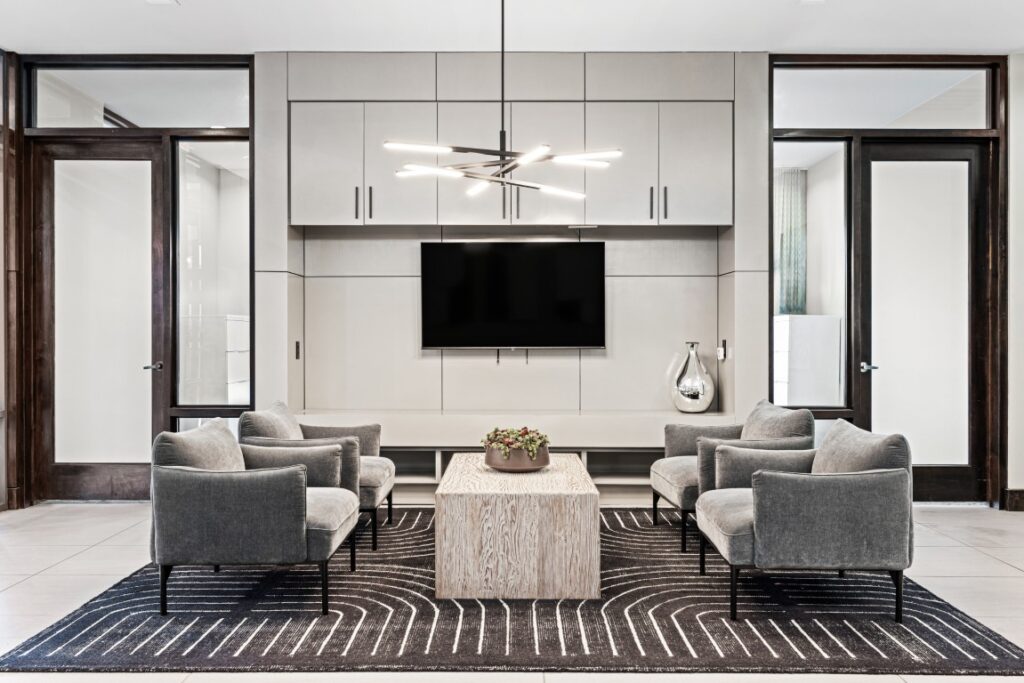 Modern lounge with four gray armchairs around a light wood coffee table, a black patterned rug, a wall-mounted TV, light cabinets, and a contemporary chandelier overhead. Minimalist decor, bright and clean.