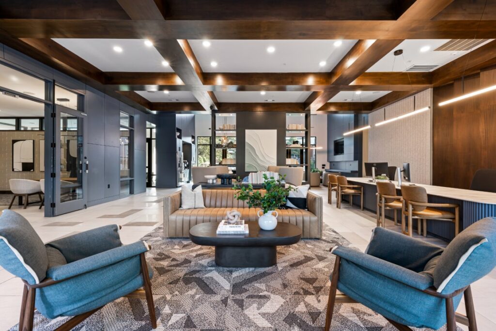 Modern office lounge with blue armchairs, tan sofa, coffee table, and geometric rug. Workstations line the right wall. Exposed wood beams and large windows create a welcoming, open atmosphere.