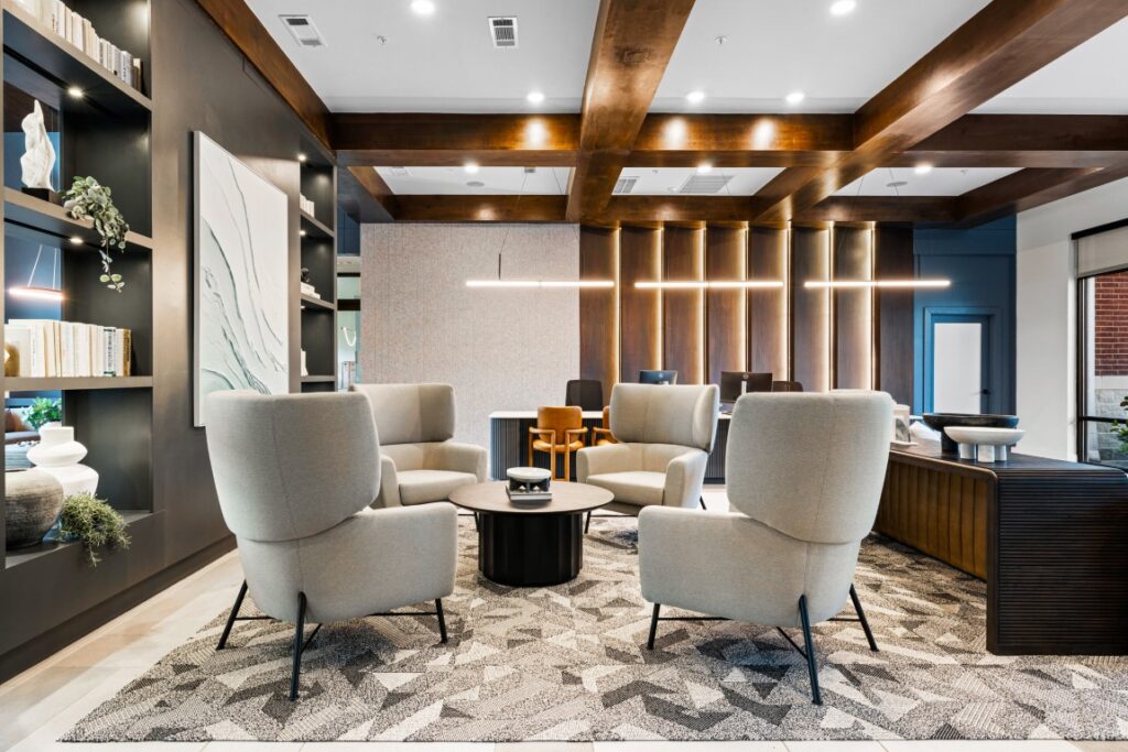 Modern lounge with four light gray armchairs around a circular coffee table, geometric rug, wooden ceiling beams, shelves with decor, abstract art, and warm lighting in a stylish, cozy setting.