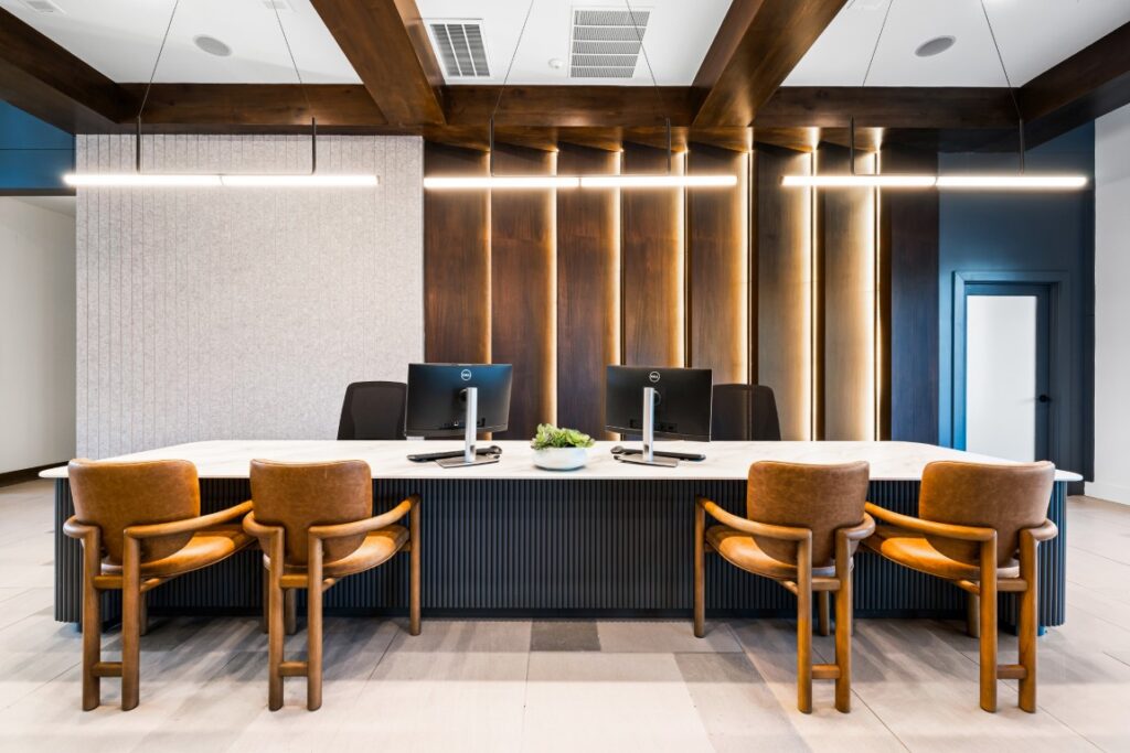 Modern office reception area with a long marble desk, two computer monitors, brown leather chairs, a small plant centerpiece, wooden beams, and vertical light accents on a wooden wall.