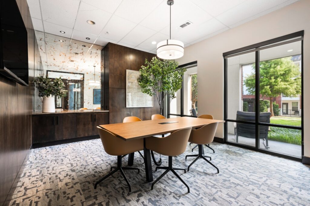 Modern conference room with wood table, four tan chairs, dark wood cabinets, large indoor plant, mirrored wall, round ceiling light, and windows showing greenery outside. Carpeted floor and neutral decor create a clean, bright space.