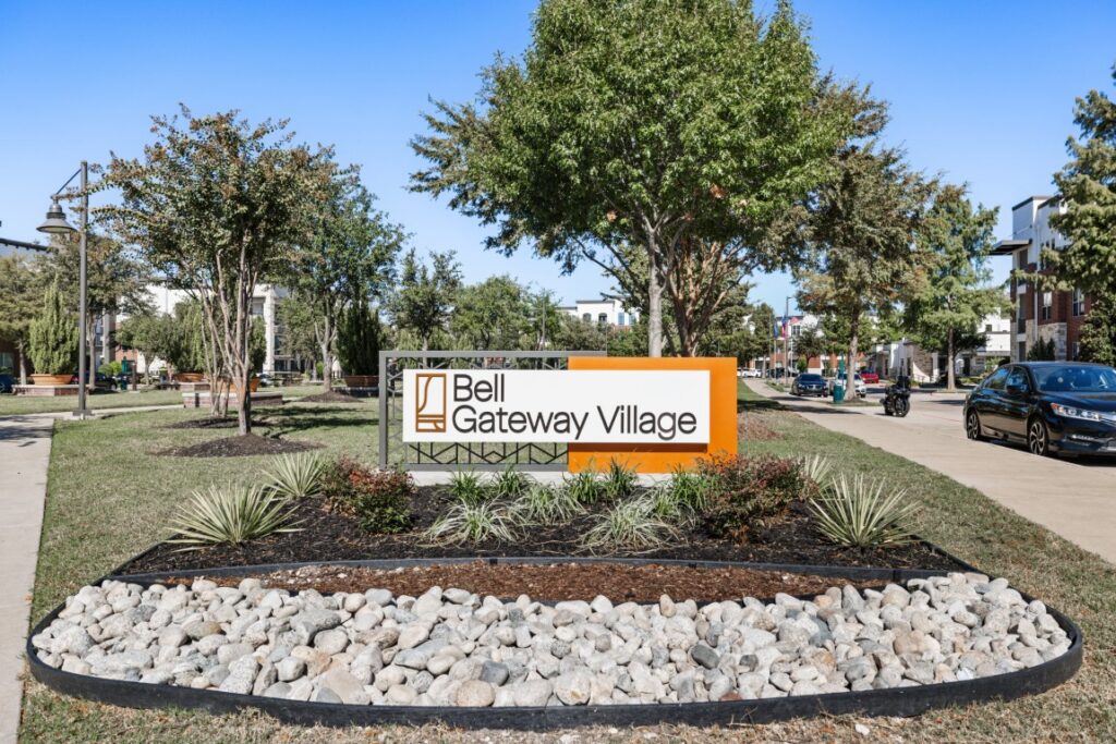 A sign reading Bell Gateway Village sits in a landscaped area with rocks and plants, surrounded by trees, modern buildings, a sidewalk, and parked cars under a clear blue sky.