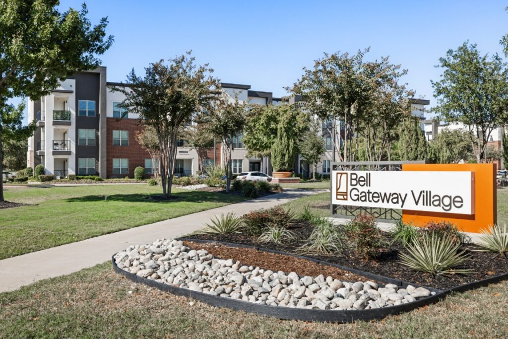 Apartment complex with three-story buildings, green lawns, and trees. In the foreground, theres a large orange and white sign that reads Bell Gateway Village. The sky is clear and blue.