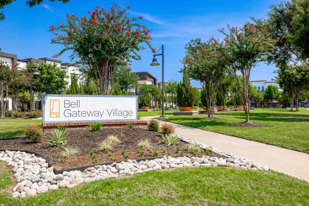 A landscaped area with a sign reading Bell Gateway Village sits beside a sidewalk, surrounded by green grass, trees, and buildings under a clear blue sky.