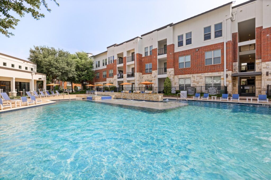 A large outdoor swimming pool with clear water, surrounded by lounge chairs and umbrellas. In the background, there is a modern three-story apartment building with red brick and white siding.