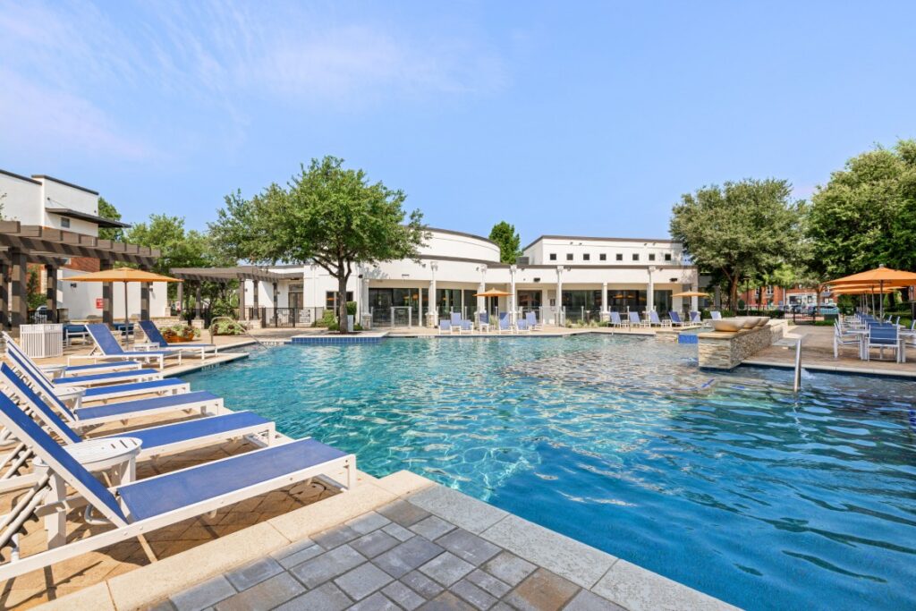 Outdoor swimming pool with blue lounge chairs, umbrellas, and a waterfall feature, surrounded by trees and white buildings under a clear sky.
