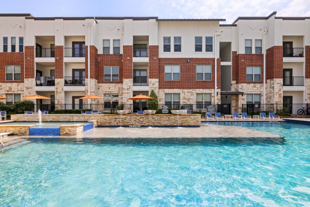 Modern apartment building with a large outdoor swimming pool in front, lounge chairs, and orange umbrellas. The building has white, brick, and stone exterior details and balconies on each level.